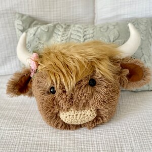 May include: A plush brown and white Highland cow head with a pink flower crown. The cow has a crocheted nose and black eyes.