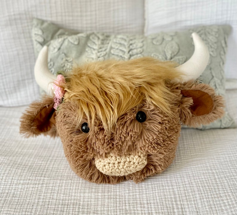 May include: A plush brown and white Highland cow head with a pink flower crown. The cow has a crocheted nose and black eyes.