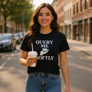 Può includere: Una maglietta nera con la scritta bianca "QUERY ME SOFTLY". La maglietta presenta una grafica di due mani che tengono un cuore. La persona indossa jeans blu e tiene in mano una tazza di caffè con una cannuccia.