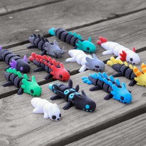 Op de afbeelding: Een verzameling kleurrijke, gestileerde axolotl-beeldjes. De beeldjes zijn in verschillende kleuren, waaronder zwart, blauw, groen, rood, wit en geel, met gedetailleerde kenmerken. Ze zijn gerangschikt op een verweerd houten oppervlak.