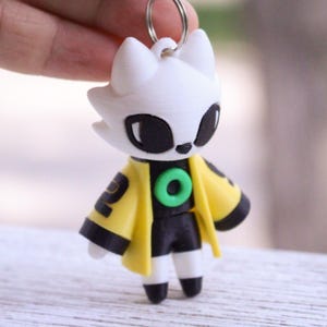 May include: A 3D printed keychain featuring a stylized white creature with large black eyes, wearing a yellow jacket with black trim and a green circular emblem on the chest. The figure has black shorts and white legs with black feet, attached to a metal ring.