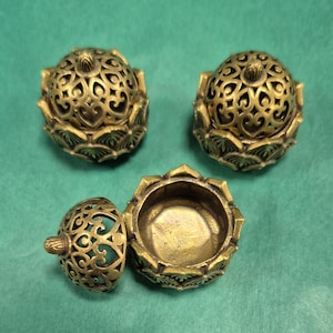 May include: Three ornate, gold-coloured incense burners with a lotus flower design. One burner is open, revealing the interior. The lids feature intricate cut-out patterns. The burners sit on a green surface.