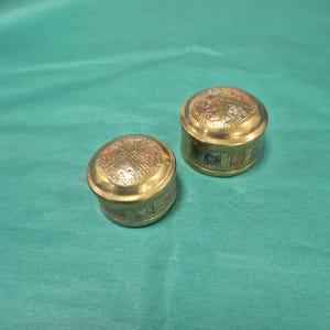 May include: Two small, round, metallic containers with lids. The containers feature a polished gold-tone finish with decorative panels on the lids and sides, displaying alternating sections of copper, silver, and gold-coloured metal with etched patterns.