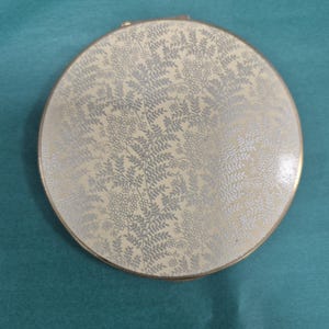 May include: A round compact mirror featuring a cream-coloured surface with a delicate, repeating silver-toned botanical pattern of small flowers and fern-like leaves. The circular case is edged with a thin metallic gold-toned rim and a small hinge.