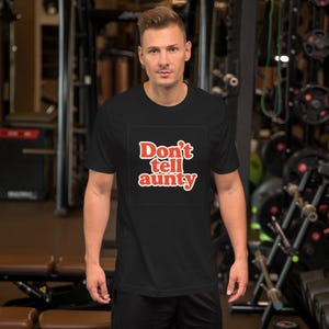 May include: Black t-shirt with the text "Don't tell aunty" in red, outlined in white. The shirt is short-sleeved and appears to be made of a soft material. The person wearing the shirt is standing in a gym.