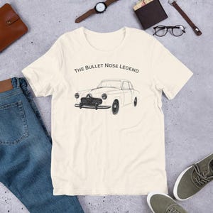 May include: A cream-coloured t-shirt with a black line drawing of a classic car. The text "THE BULLET NOSE LEGEND" is printed above the car. The shirt is made of soft material and is suitable for casual wear.