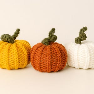 May include: Three knitted pumpkins in fall colors. One is yellow, one is orange, and one is white. Each pumpkin has a green stem. The pumpkins are arranged side by side, perfect for autumn decor.