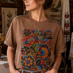 May include: A brown t-shirt featuring a vibrant floral design in red, blue, yellow, and green. The shirt has short sleeves and is paired with blue jeans and a brown belt. The background shows a rustic interior with wooden walls and traditional decor.