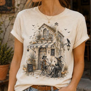 May include: A cream-colored t-shirt featuring a graphic illustration of a skeleton and a witch sitting at a cafe table outside a building. The design includes pumpkins, a crow, bats, and a sign that reads Pumpkin Season, all in a muted color palette.