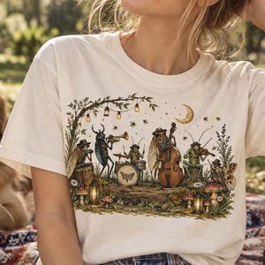 May include: An off-white short-sleeve t-shirt featuring a detailed graphic print of insects playing musical instruments in a forest setting with mushrooms, lanterns, and a crescent moon. The design includes a beetle, cicada, and grasshopper.