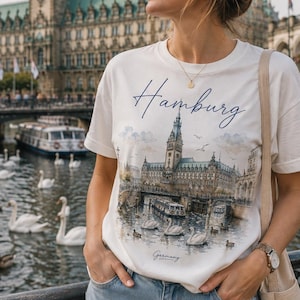 Peut inclure: Un t-shirt blanc à manches courtes orné d'une impression graphique de l'hôtel de ville de Hambourg, d'un canal avec des cygnes, et du texte Hamburg et Germany en écriture cursive. Le t-shirt a un col rond et une coupe décontractée.