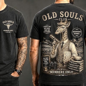 May include: A black t-shirt featuring a small graphic on the front chest with a bull skull and the text OLD SOULS CLUB MEMBERS ONLY EST 2025. The back displays a giraffe in a vest holding a cigar with text including OLD SOULS CLUB.