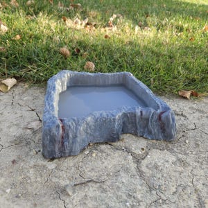 Może przedstawiać: Szara, prostokątna miska na wodę dla zwierząt domowych z teksturowanym, skalistym wyglądem zewnętrznym. Wnętrze zawiera jasno szarą ciecz. Miska stoi na spękanej, suchej ziemi, z trawą i opadłymi liśćmi w tle.