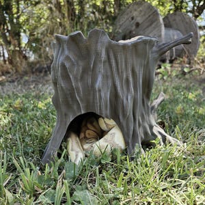 Op de afbeelding: Een grijs, boomstronk-vormig reptielenverblijf met een uitgehold midden. Een slang is opgerold in het verblijf. Het verblijf staat in een grasveld.