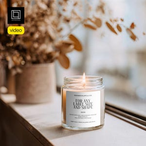 May include: A burning candle in a clear glass jar with a white label. The label states "FOR ANY LABEL SIZE AND SHAPE" and "250g". The candle is on a wooden surface, with dried flowers in the background.