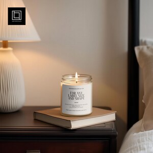 May include: A lit candle in a clear glass jar with a white label that reads "FOR ANY LABEL SIZE AND SHAPE" sits on a closed book. The candle is on a dark wooden nightstand next to a lamp.