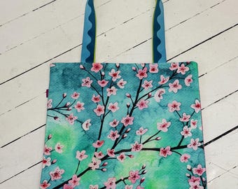 Könnte beinhalten: Eine quadratische Tragetasche mit einem Aquarell-Druck von rosa Kirschblüten auf dunklen Zweigen vor einem türkisfarbenen und grünen Hintergrund. Die Tasche hat zwei blaue Schulterriemen mit einem dunkelblauen Wellenmuster und einem lindgrünen Rand.