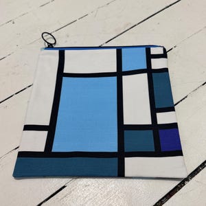 May include: A rectangular canvas pouch with a Mondrian-style geometric design. The design features a grid of black lines and blocks of white, light blue, and dark blue. The pouch has a blue zipper and a black zipper pull.