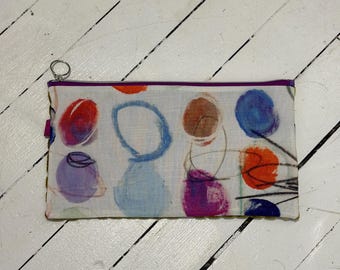 Puede incluir: Un estuche rectangular con cremallera sobre una base de tela blanca, decorado con un patrón abstracto de círculos dibujados a mano en tonos rojo, azul, morado y naranja, acentuado por finas líneas negras y una cremallera morada con anilla.