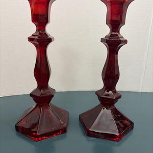 May include: A pair of matching ruby red glass candlesticks featuring a faceted hexagonal base, a slender stem with a central bulbous section, and a flared top rim designed to hold a standard taper candle securely in place.