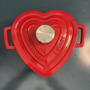 Puede incluir: Una olla de hierro fundido esmaltado de color rojo brillante en forma de corazón con dos asas laterales y un pomo de metal plateado en la tapa. La tapa presenta el texto en relieve MARTHA STEWART y 2 QUART. La olla tiene un acabado brillante.