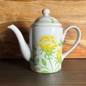 May include: White ceramic teapot with a lid, featuring a yellow rose floral design with green leaves and stems. The teapot has a curved spout and a handle. The background is a wooden surface.