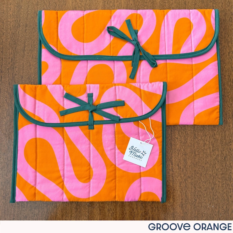 May include: Two quilted fabric cases in orange and pink with a wavy pattern. The cases have a dark green trim and a matching bow closure. A white tag with the text "Hello Meko Handmade" is attached to the smaller case.