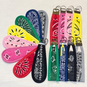 May include: A collection of keychains and pen holders in various colors, featuring a paisley bandana pattern. The keychains have metal rings and are in shades of red, pink, yellow, green, blue, and black. The pen holders are designed to hold pens and other small items.