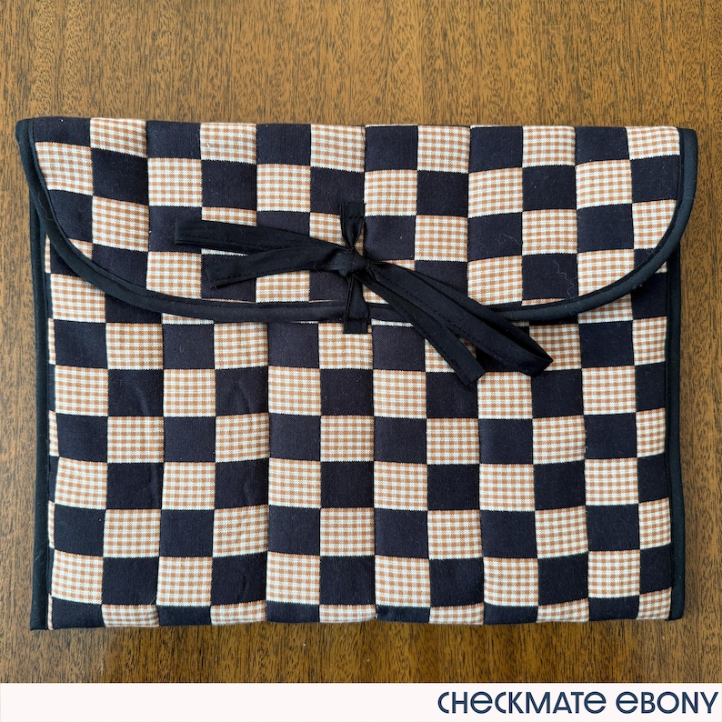 May include: A black, tan, and white checkered fabric case with a black trim and a black tie closure. The case is rectangular with rounded corners and the text "CHECKMATE EBONY" is at the bottom.
