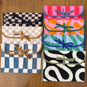 May include: A collection of colorful, quilted fabric pouches with curved flap closures. The pouches feature various patterns, including checkerboard, stripes, and abstract designs. Each pouch is secured with a matching fabric tie, offering a stylish and functional storage solution.