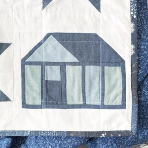May include: Close-up of a handmade quilt with a house applique. The quilt features a white background with blue and light blue house and triangle shapes. The edges are trimmed with a blue floral pattern.