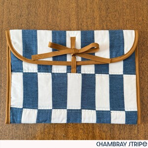 May include: A blue and white striped fabric case with a brown leather trim and bow closure. The case has a curved flap and the text "CHAMBRAY STRIPE" at the bottom.