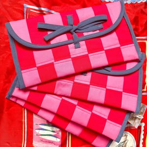 May include: Two red and pink checkered fabric pouches with gray trim and a bow closure. The pouches are stacked on a red background with food-themed illustrations.