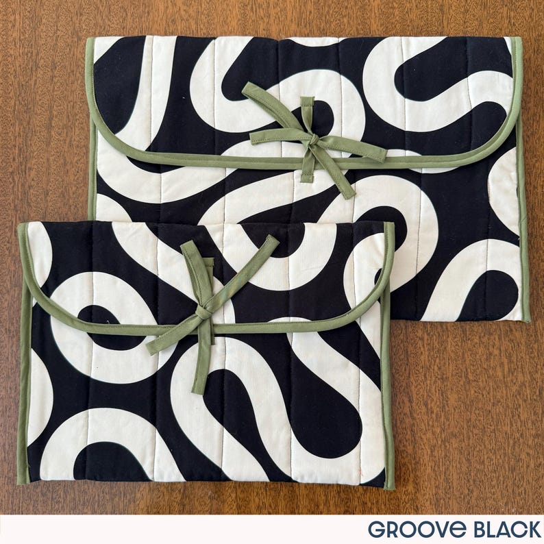 May include: Two black and white quilted fabric cases with a wavy pattern. The cases have a rounded flap with a green trim and a matching tie closure. The cases are in different sizes and are on a wooden surface. The text "GROOVE BLACK" is at the bottom.