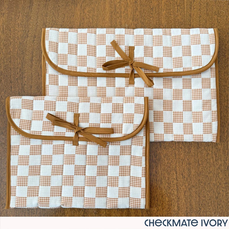 May include: Two ivory and brown checkered fabric pouches with rounded edges and brown ribbon ties. The pouches are different sizes and are made of a quilted material. The text "CHECKMATE IVORY" is at the bottom.