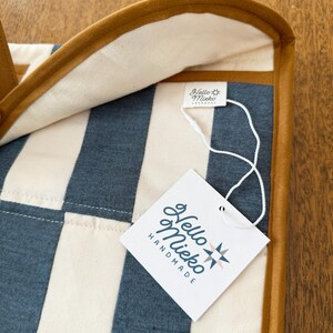 May include: Close-up of a handmade item with a striped pattern in blue and cream. The item has a brown leather trim and a tag that reads "Hello Mieko Handmade". The background is a wooden surface.