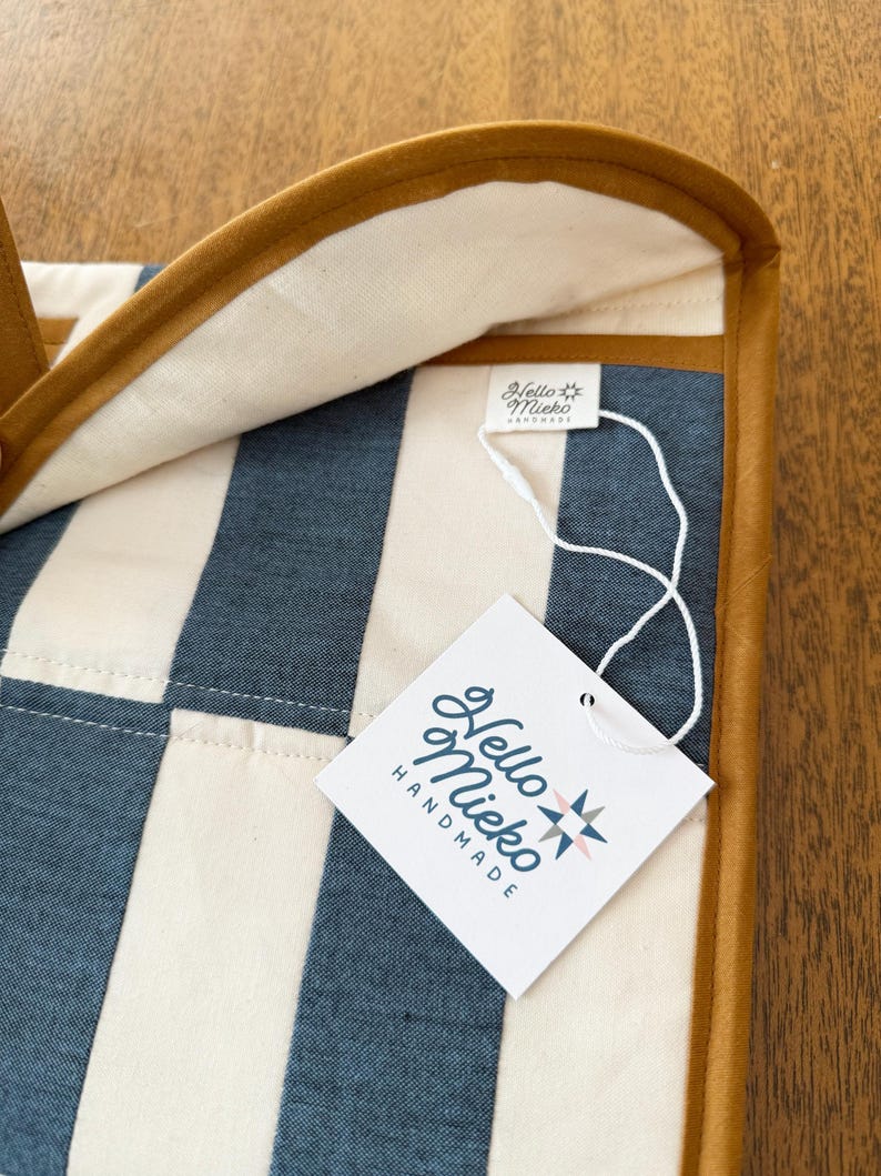 May include: Close-up of a handmade item with a striped pattern in blue and cream. The item has a brown leather trim and a tag that reads "Hello Mieko Handmade". The background is a wooden surface.