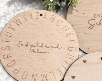 Könnte beinhalten: Drei kreisförmige, helle Holzscheiben mit eingravierten Details. Eine Scheibe zeigt das Alphabet im Kreis um den mittigen Text "Schulkind Malou". Weitere Scheiben zeigen den Teiltext "END" und "Sch" mit kleinen eingravierten Herzen.