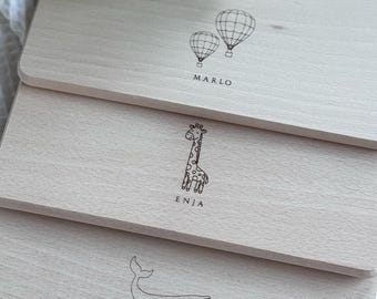 Könnte beinhalten: Drei hellfarbene Holzschneidebretter mit eingravierten Designs. Das obere Brett zeigt zwei Heißluftballons und den Namen "MARLO". Das mittlere Brett hat eine Giraffe und den Namen "ENJA". Das untere Brett zeigt einen Wal und den Namen "MAJALENA".