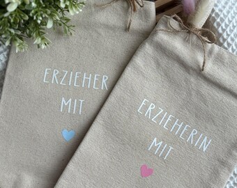 Könnte beinhalten: Zwei beigefarbene Tragetaschen mit weißem Text und Herzgrafiken. Auf einer Tasche steht "ERZIEHER MIT" mit einem blauen Herzen, auf der anderen "ERZIEHERIN MIT" mit einem rosa Herzen. Jede Tasche hat einen dekorativen floralen Akzent.