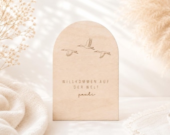 Könnte beinhalten: Eine bogenförmige Platte aus hellem Holz mit einer Gravur von drei fliegenden Vögeln. Der Text "WILLKOMMEN AUF DER WELT" ist in Großbuchstaben über dem Namen "pauli" eingraviert, der in einer kursiven Schriftart dargestellt ist.