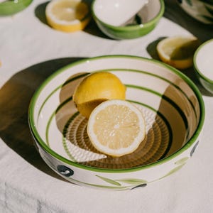 May include: A ceramic grater bowl with a cream-coloured interior featuring a textured centre for grating and green painted stripes. The exterior is decorated with green leaves and dark blue olives. A whole lemon and a half lemon sit inside the bowl.