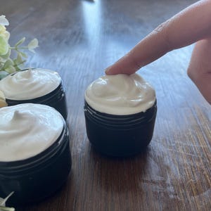 May include: Three small, black jars filled with a white, whipped cream-like substance. A finger gently touches the top of one jar. The jars are on a wooden surface, with flowers in the background.