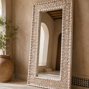 Peut inclure: Un grand miroir rectangulaire avec un cadre en bois sculpté blanc cassé. Le cadre présente des motifs géométriques complexes. Le miroir est adossé à un mur beige dans une pièce avec de la lumière naturelle et un pot en terre cuite avec une plante.