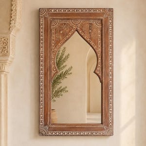 Puede incluir: Espejo rectangular de madera marrón con parte superior arqueada. El marco presenta intrincados patrones florales y geométricos tallados con detalles en blanco. El espejo refleja una habitación con una planta y una puerta. Decoración de pared.