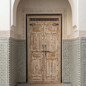 May include: A weathered, light-colored wooden double door with metal hardware, set within an arched entryway. The door features rectangular panels and decorative metal studs. The surrounding walls are adorned with patterned tiles and ornate carvings, creating a rustic, yet elegant aesthetic.