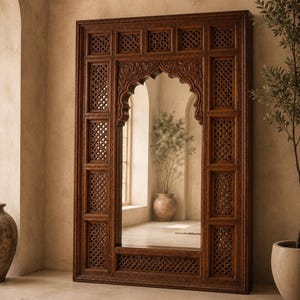 May include: A large, rectangular wooden mirror with intricate carved details. The frame is a rich brown color and features an arched top and lattice-like side panels. The mirror reflects a room with natural light and potted plants, creating a warm, inviting atmosphere.