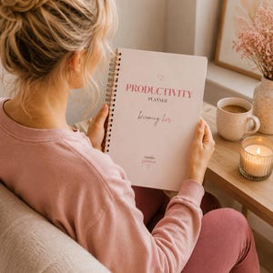 Op de afbeelding: Een roze Productiviteitsplanner met een spiraalbinding is opengeklapt. De cover leest "PRODUCTIVITY PLANNER" en "becoming her" in roze script. Een brandende kaars, een mok koffie en een ingelijste foto staan op een houten tafel.