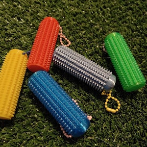 Op de afbeelding: Een verzameling kleurrijke, cilindervormige hondenspeeltjes in geel, rood, blauw, groen en zilver. Elk speeltje heeft een getextureerd oppervlak met kleine, puntige uitsteeksels. Ze zijn gerangschikt op een groene, kunstgras ondergrond.