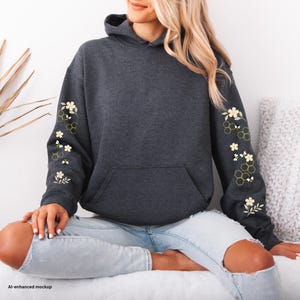 May include: A dark charcoal grey hooded sweatshirt featuring a front kangaroo pocket and long sleeves decorated with a repeating pattern of small white flowers, green leaves, yellow honeycombs, and tiny black and yellow bees on both arms.
